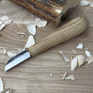 May include: A close-up of a wood carving knife with a wooden handle and a sharp blade. The knife is lying on a wooden surface with wood shavings around it.