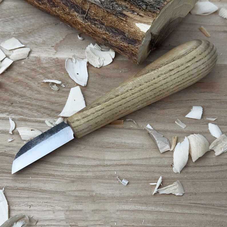 May include: A close-up of a wood carving knife with a wooden handle and a sharp blade. The knife is lying on a wooden surface with wood shavings around it.
