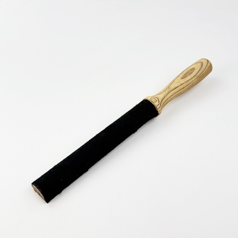May include: A long, cylindrical tool featuring a black, textured, felt-like covering on the main body and a light-colored wooden handle with a visible grain pattern. The handle is shaped for a grip, transitioning into the straight black section.