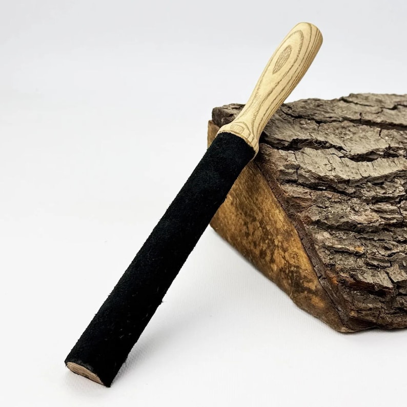 May include: A long, cylindrical tool featuring a light-colored, natural wood handle with visible grain patterns and a long, black, textured suede-like material covering the main body of the shaft, presented at an angle against a white background.