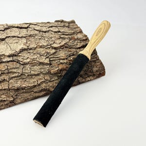 May include: A sharpening strop featuring a long, rectangular black leather surface attached to a light-colored wooden handle with a visible grain pattern. The tool is designed for honing blades and has a smooth, finished wooden grip.