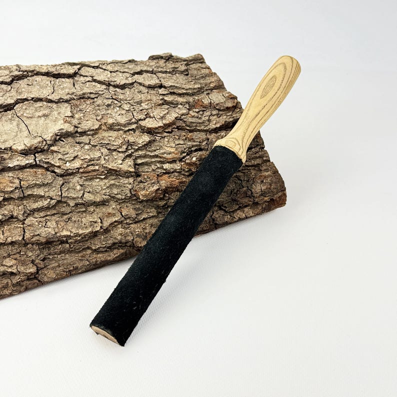May include: A sharpening strop featuring a long, rectangular black leather surface attached to a light-colored wooden handle with a visible grain pattern. The tool is designed for honing blades and has a smooth, finished wooden grip.
