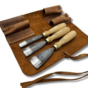 以下が含まれることがあります： 金属刃と木製ハンドルを備えた3つの木彫りノミが、茶色の革製ツールロールに収められています。ノミは一列に並べられ、鋭い刃先と職人技が際立っています。ツールロールは紐で閉じるタイプです。