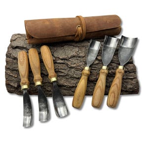 以下が含まれることがあります： 木製のハンドルと金属製の刃が付いた6つの木彫り工具のセット。茶色の革製ツールロールと一緒に展示されています。ツールには真鍮のアクセントがあり、質感のある樹皮の上に配置されています。