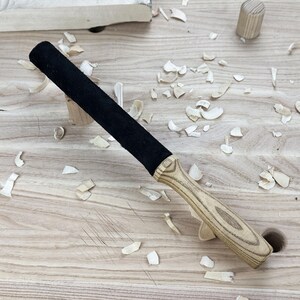May include: A leather honing strop featuring a long, rectangular black leather surface attached to a light-colored, grained wooden handle. The tool is designed for sharpening blades and is shown with a smooth, ergonomic grip and a sturdy construction.