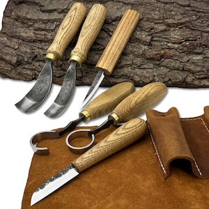 May include: A set of wood carving tools with wooden handles and metal blades. The tools include curved gouges, a straight knife, and a hook knife, displayed on a brown leather surface. A leather sheath is also included.