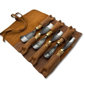 May include: A set of six wood carving gouges with wooden handles and metal blades, presented in an open brown leather tool roll. The roll has a leather tie closure.