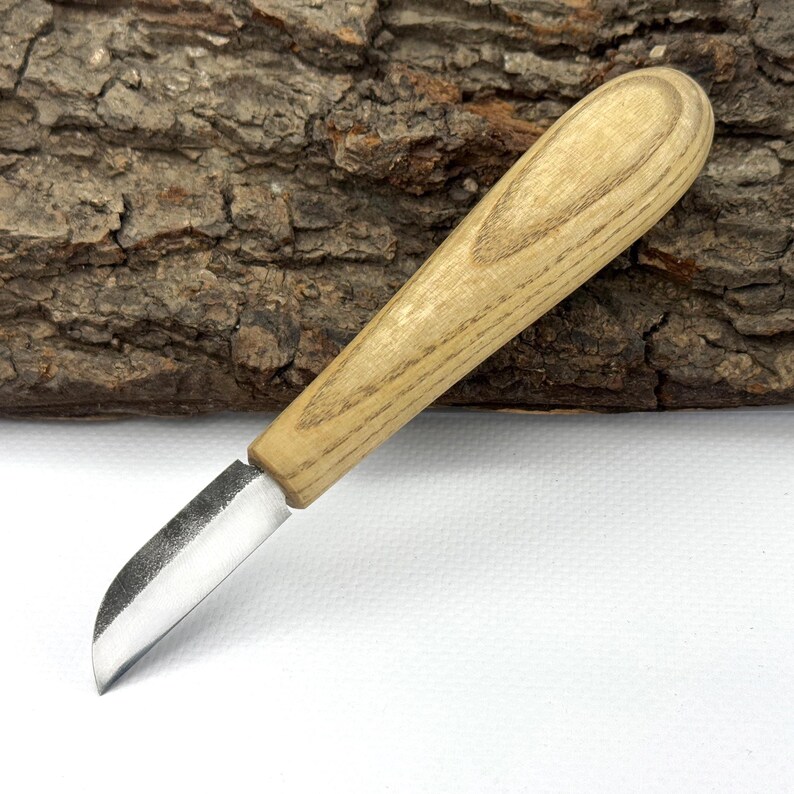 May include: A small, hand-crafted wood carving knife with a curved blade and a wooden handle. The knife is resting on a piece of tree bark.