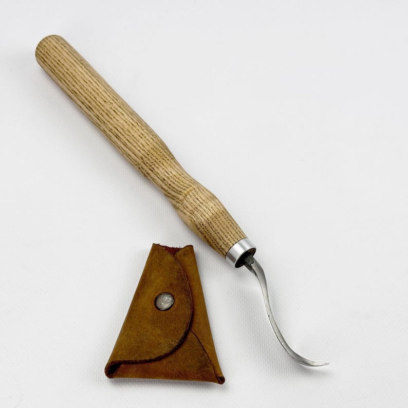 May include: A wooden handled carving tool with a curved blade and a brown leather sheath. The tool is designed for carving wood.