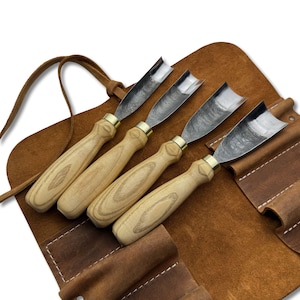 May include: Four wood carving gouges with light wood handles, brass ferrules, and curved steel blades. They are presented in a brown leather tool roll with a leather strap for storage.