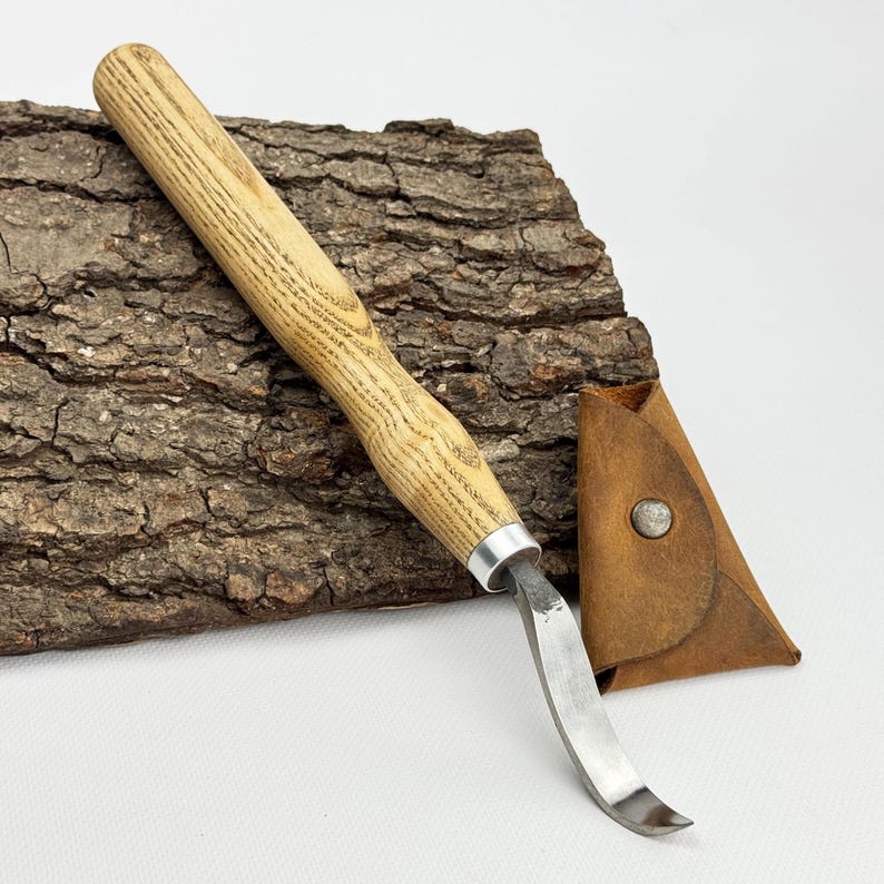May include: A wooden handled carving knife with a curved blade and a leather sheath. The knife is resting on a piece of bark.