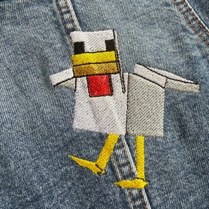 May include: A white chicken with yellow feet and a red beak is embroidered on a blue denim jacket. The chicken is wearing a white shirt.