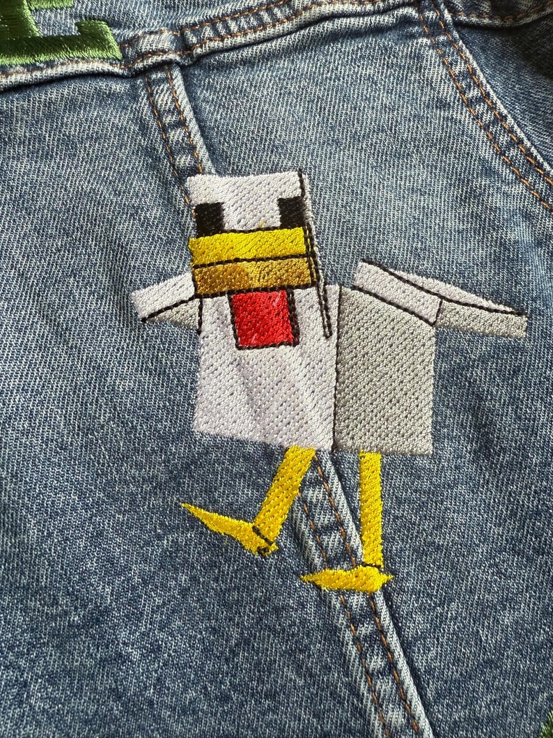 May include: A white chicken with yellow feet and a red beak is embroidered on a blue denim jacket. The chicken is wearing a white shirt.