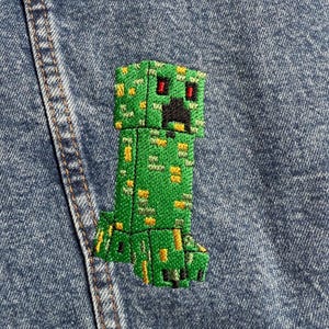 May include: Green Minecraft Creeper embroidered patch with red eyes and black mouth on a blue denim background.