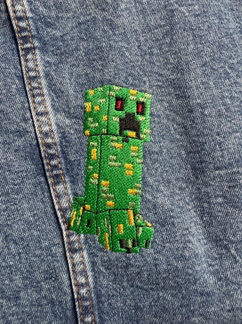 May include: Green Minecraft Creeper embroidered patch with red eyes and black mouth on a blue denim background.