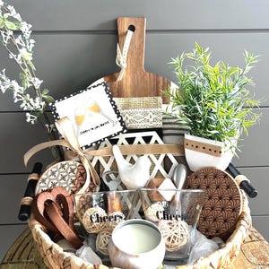 Pode incluir: Uma cesta de presente com dois copos "Cheers", uma vela e itens decorativos, apresentada em uma tábua de corte de madeira. A cesta inclui uma pequena planta, porta-copos e um cartão com o texto "Cheers to you both".