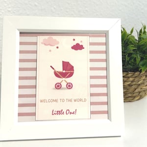 May include: A square white picture frame containing a print with a pink baby carriage illustration, pink clouds, and the text "WELCOME TO THE WORLD Little One!" set against a background of horizontal pink and white stripes.