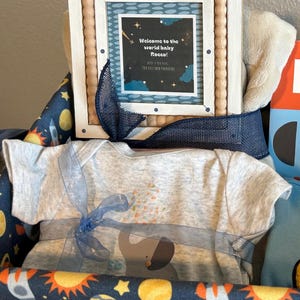 May include: A baby gift basket with a gray onesie featuring an elephant design, tied with a blue ribbon. The basket includes a framed card that reads "Welcome to the world baby Rocco!" and is decorated with a space-themed fabric.