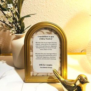 May include: An arched gold picture frame containing a printed message that reads: Congratulations on your wedding Christina! We pray that as you begin this new chapter, Jesus will be the center of your home. With love & prayers. Your Mentor Group.