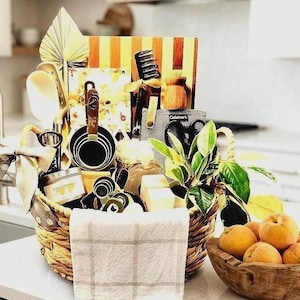May include: A woven basket filled with kitchen essentials including a striped wooden cutting board, a set of black measuring cups, a white spatula, a small candle, a grey Cuisinart oven mitt, a pair of black scissors, and a small green plant.