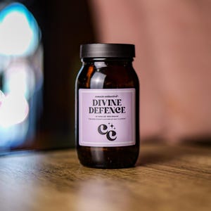 Könnte beinhalten: Eine braune Glasflasche mit schwarzem Deckel und einem lilafarbenen Etikett, auf dem "cosmic collective" und "DIVINE DEFENCE 60 KAPSELN 100% BIO FÜR HAARSTÄRKE UND UNTERSTÜTZUNG DER IMMUNGESUNDHEIT" steht. Das Etikett zeigt auch ein stilisiertes "CC"-Logo mit Sternen.