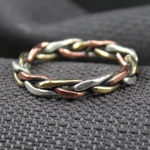 May include: A braided ring with silver, gold, and copper-colored metal strands. The ring is set against a dark gray textured fabric background. The ring's design features an interwoven pattern.