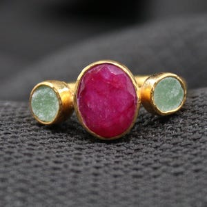 May include: A gold ring featuring a large oval ruby center stone flanked by two smaller, round, light green stones. The ring is set against a dark gray textured background.