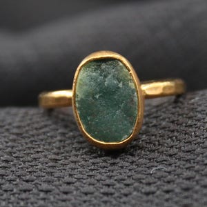 May include: A gold ring featuring an oval, rough-cut, green gemstone. The ring has a simple band and a bezel setting. The gemstone has a textured appearance. The ring is displayed on a dark gray fabric.
