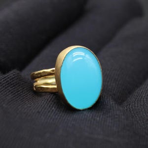 May include: A gold-colored ring with a large, oval, light blue stone. The ring has a double band design and a smooth, polished finish. The stone is set in a gold bezel.
