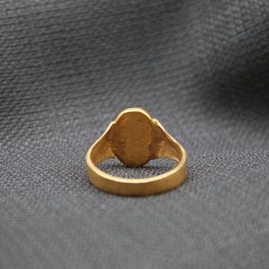 May include: A gold ring with a smooth, oval-shaped stone set in the center. The ring is on a gray fabric background.