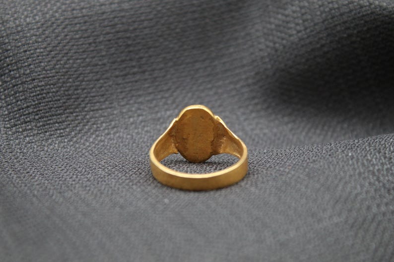 May include: A gold ring with a smooth, oval-shaped stone set in the center. The ring is on a gray fabric background.