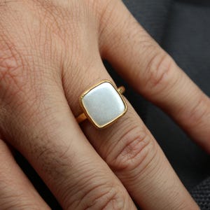 May include: A gold ring with a square, light-colored stone set in a gold band. The ring is on a person's finger, and the background is a dark gray color.