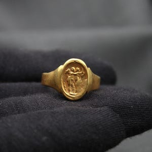 May include: A gold ring with an oval-shaped bezel featuring a raised design of two figures holding hands.
