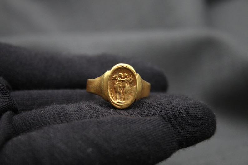 May include: A gold ring with an oval-shaped bezel featuring a raised design of two figures holding hands.