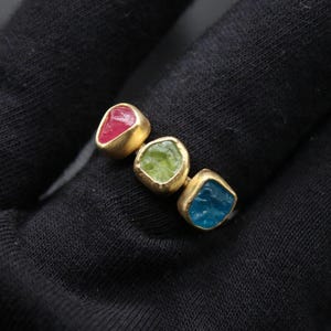 May include: A gold ring set with three faceted gemstones. The gemstones are red, green, and blue.