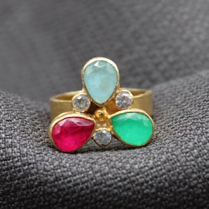 May include: A gold ring with a cluster of three teardrop-shaped gemstones. The gemstones are a light blue, a deep green, and a bright red.