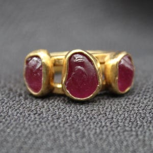 May include: Gold ring featuring three red gemstone settings. The gemstones are oval shaped and have a slightly irregular cut.
