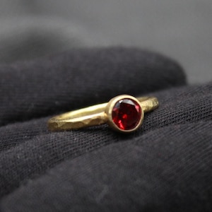 May include: A gold ring with a red gemstone set in the center. The ring has a hammered texture.