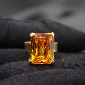 Op de afbeelding: Een gouden ring met een grote, rechthoekige, oranje-amberkleurige edelsteen.