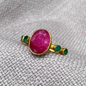 May include: A gold ring featuring a large, oval-shaped, deep pink ruby set in the center. The band is accented with two small, round, vibrant green emeralds on each side of the central stone, creating a symmetrical design with a polished finish.