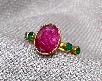May include: A gold ring featuring a large, oval-shaped, deep pink ruby set in the center. The band is accented with two small, round, vibrant green emeralds on each side of the central stone, creating a symmetrical design with a polished finish.