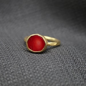 May include: A gold ring with a red gemstone set in a textured bezel. The ring is on a gray fabric background.