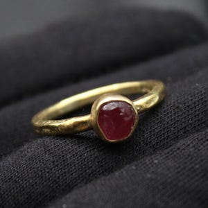 May include: A gold ring with a textured band and a red gemstone set in the center.