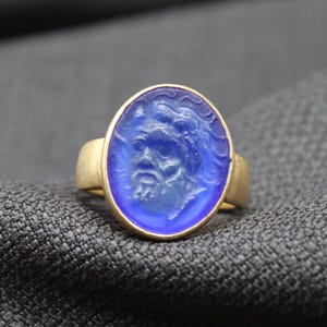May include: A gold ring with a blue oval-shaped stone featuring a carved face. The stone is set in a gold band.