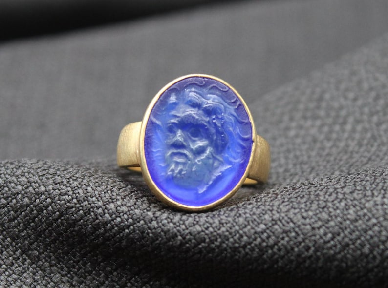 May include: A gold ring with a blue oval-shaped stone featuring a carved face. The stone is set in a gold band.