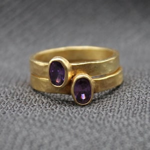 May include: Two gold rings with purple oval gemstones. The rings are stacked on top of each other.