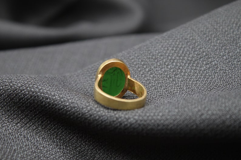 May include: A gold ring with a green oval gemstone. The ring is on a gray fabric background.