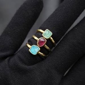 Può includere: Tre anelli d'oro con pietre preziose di diversi colori. Gli anelli sono impilati uno sopra l'altro. L'anello superiore ha una pietra preziosa verde chiaro, l'anello centrale ha una pietra preziosa rosa e l'anello inferiore ha una pietra preziosa blu chiaro.