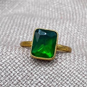 May include: A gold-toned ring featuring a large, rectangular, emerald-green faceted gemstone set in a simple metal band. The stone displays deep green hues with light reflections on its surface, while the band has a textured, matte finish.
