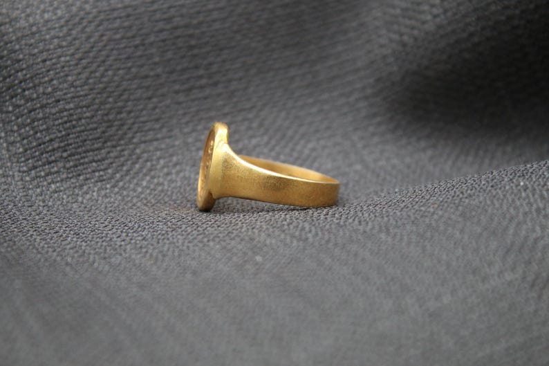 May include: A gold signet ring with a smooth, oval-shaped top. The ring is shown from the side, with the band facing the viewer.